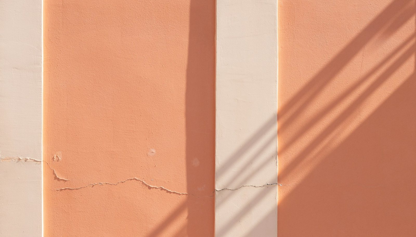 A peach stucco wall with white columns, long afternoon shadows, and a thin crack running across it.